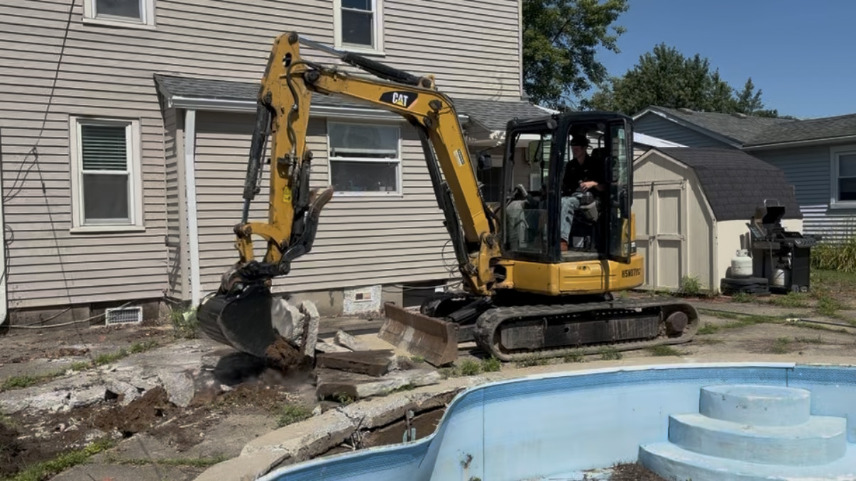 Excavator breaking up the concrete decking around a backyard swimming pool